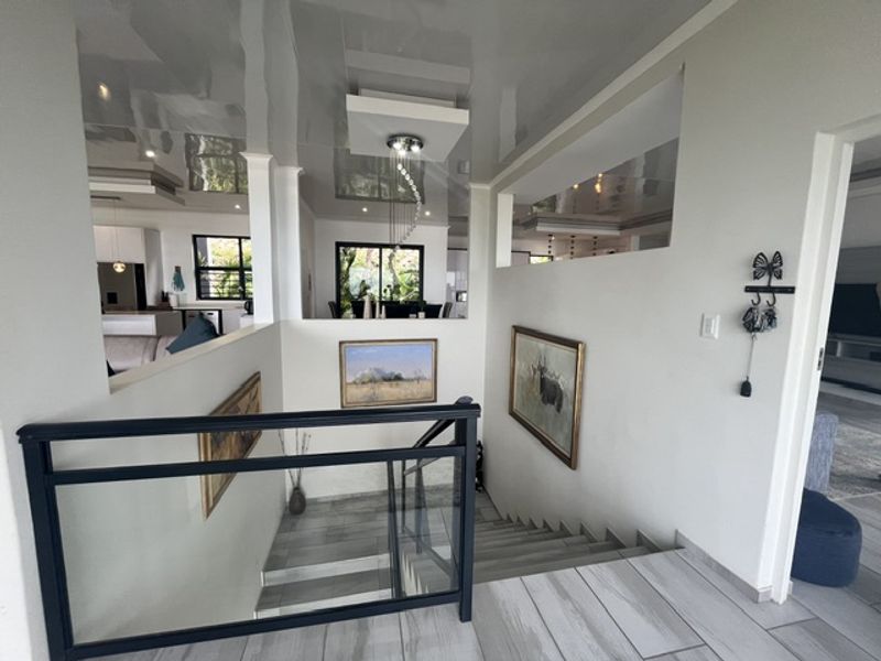 A modern hallway featuring a glass balustrade staircase, glossy ceiling finish, and neutral tiled flooring, providing an open view into the adjacent living areas.