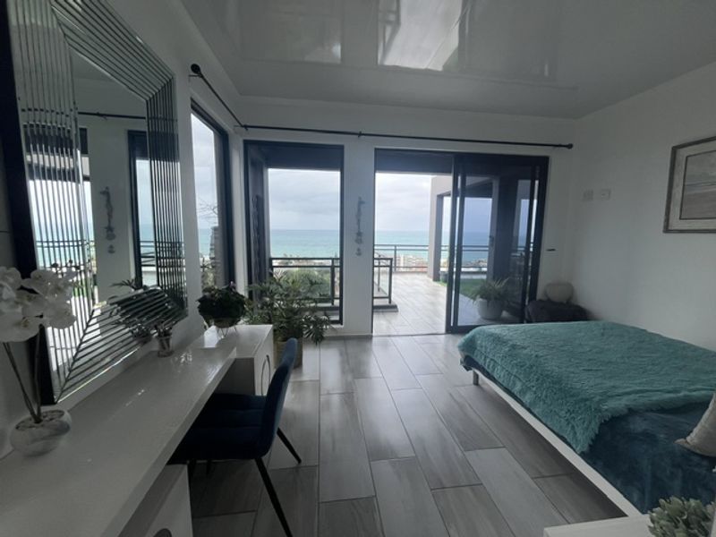 Modern bedroom featuring glossy ceiling, tiled floors, and large sliding glass doors opening onto a balcony with scenic ocean views.