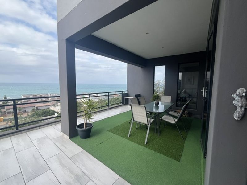 A spacious covered balcony featuring modern tiled flooring, artificial grass section, and a metal balustrade with expansive ocean views.