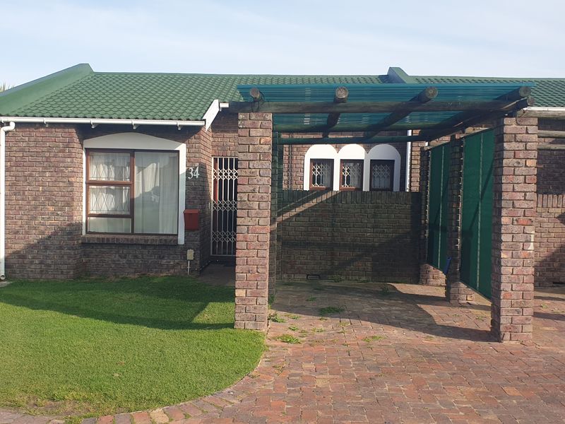 Front elevation of a facebrick townhouse featuring a green tiled roof, a paved driveway with a pergola carport, and a well-maintained front lawn.