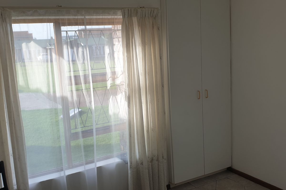 A bright, empty bedroom featuring built-in cupboards, tiled flooring, and a large window with security bars overlooking the garden.