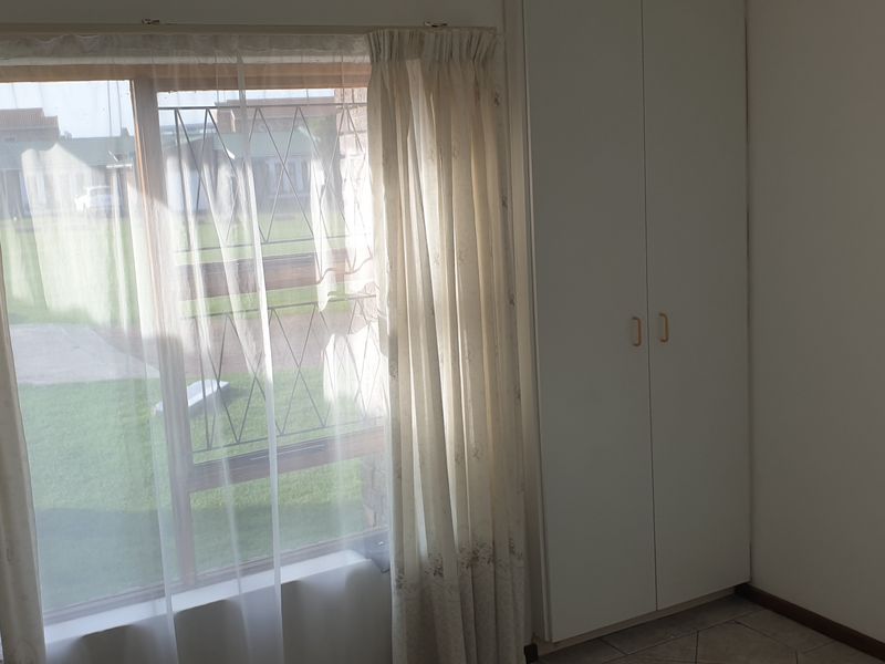 A bright, empty bedroom featuring built-in cupboards, tiled flooring, and a large window with security bars overlooking the garden.