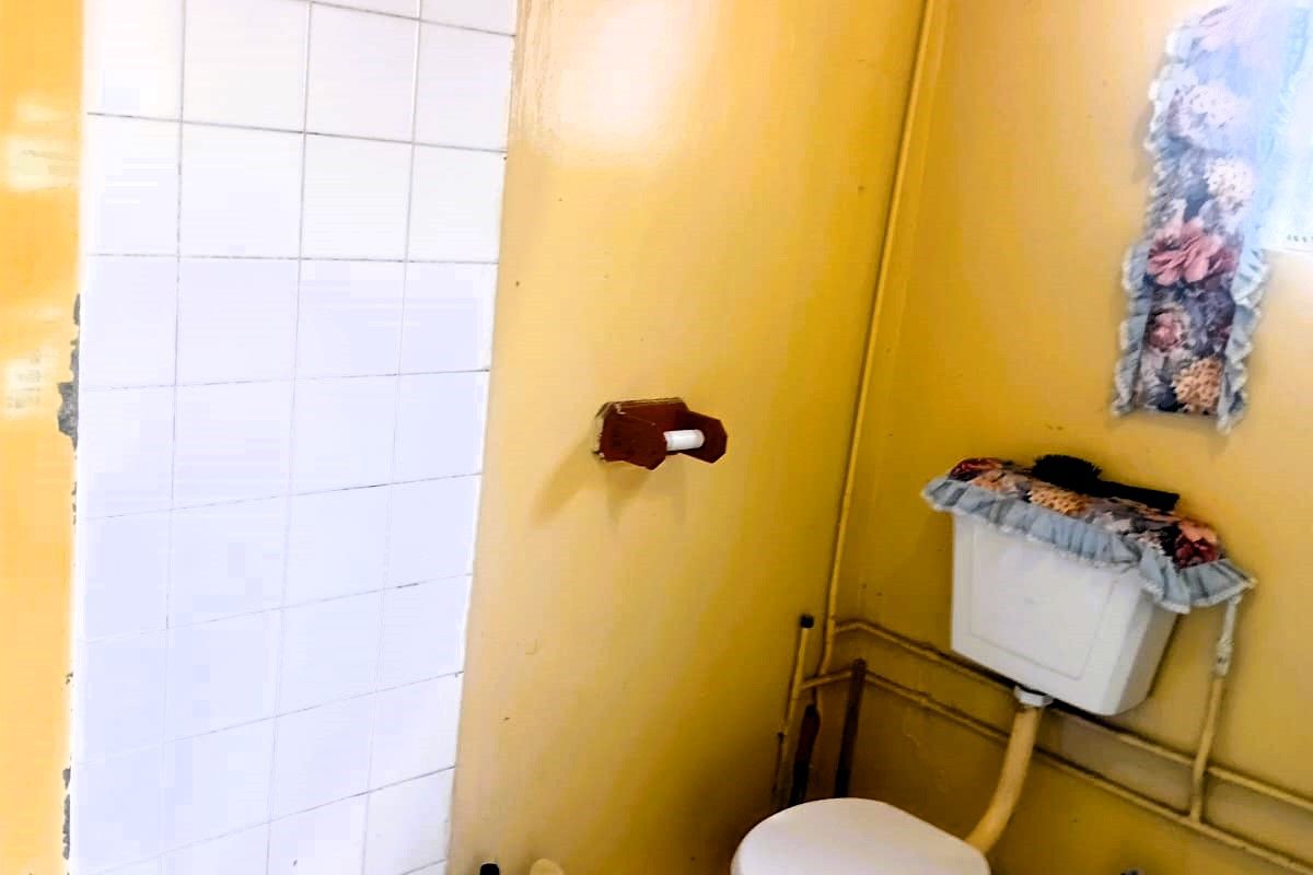 A dated bathroom featuring a toilet and tiled shower area, currently showing significant wear and tear on the walls and finishes.