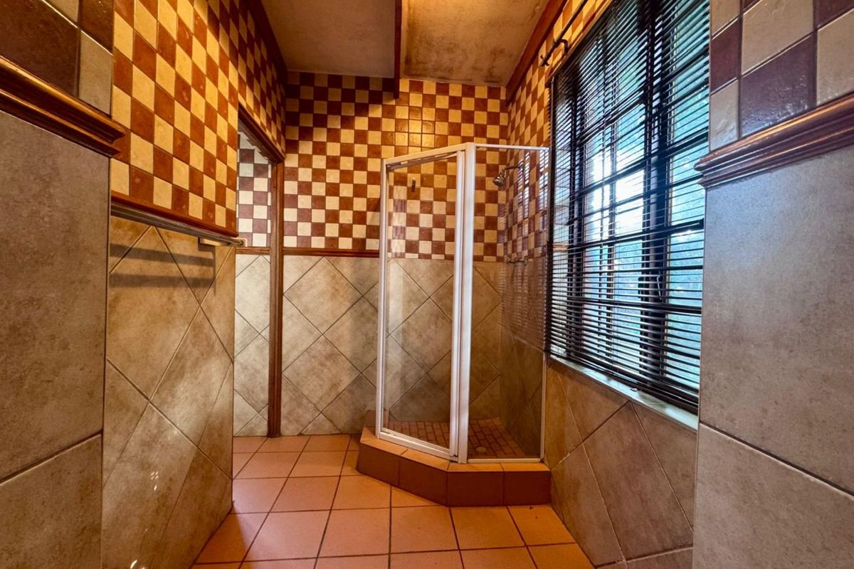 A dated bathroom featuring a corner shower enclosure, tiled walls with a checkered pattern, and a window with fitted blinds.