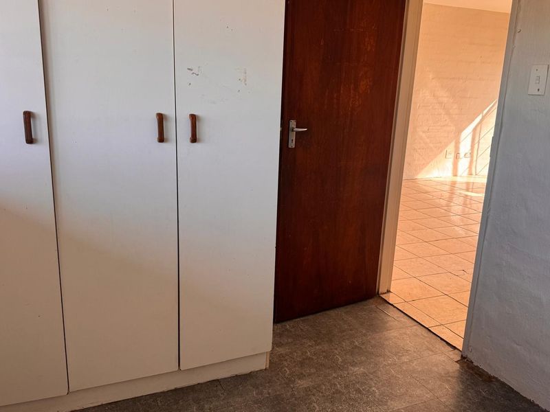 A bedroom interior featuring built-in cupboards and a wooden door, leading into an adjacent tiled living space.