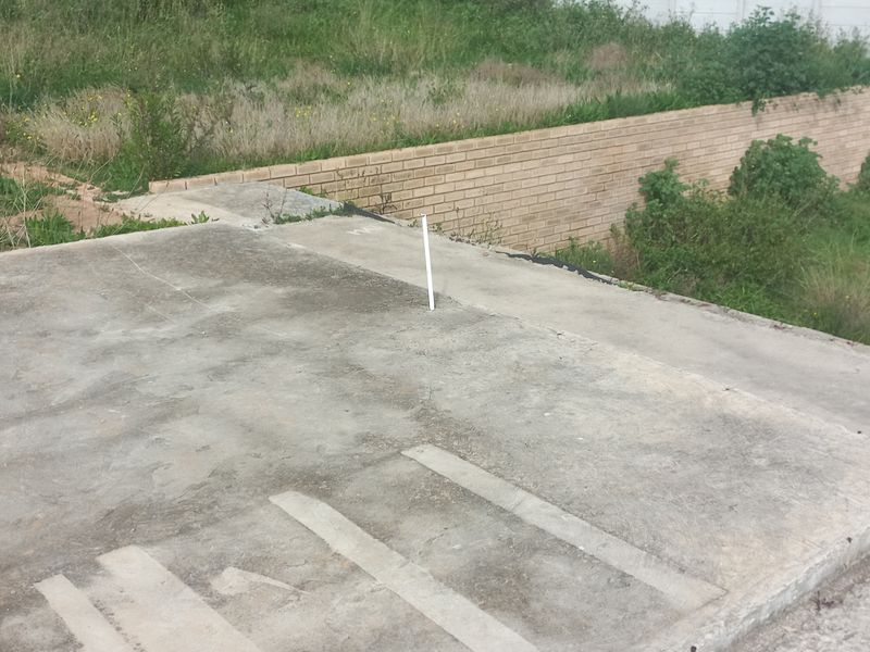 A view of a vacant residential plot featuring a concrete slab foundation, a brick retaining wall, and surrounding overgrown vegetation.