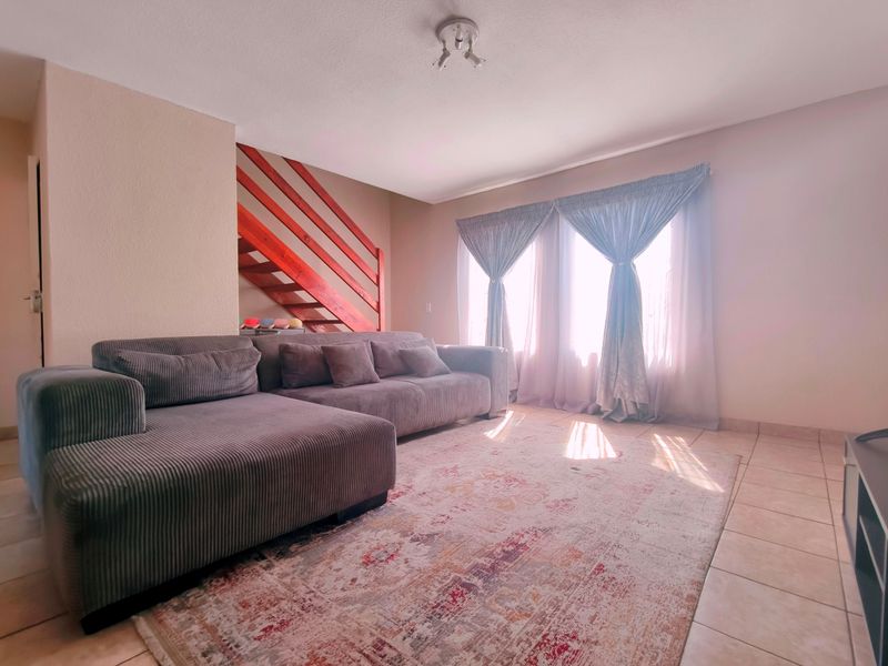 A bright living room featuring tiled flooring and a prominent wooden staircase with a balustrade, offering a functional and open layout.