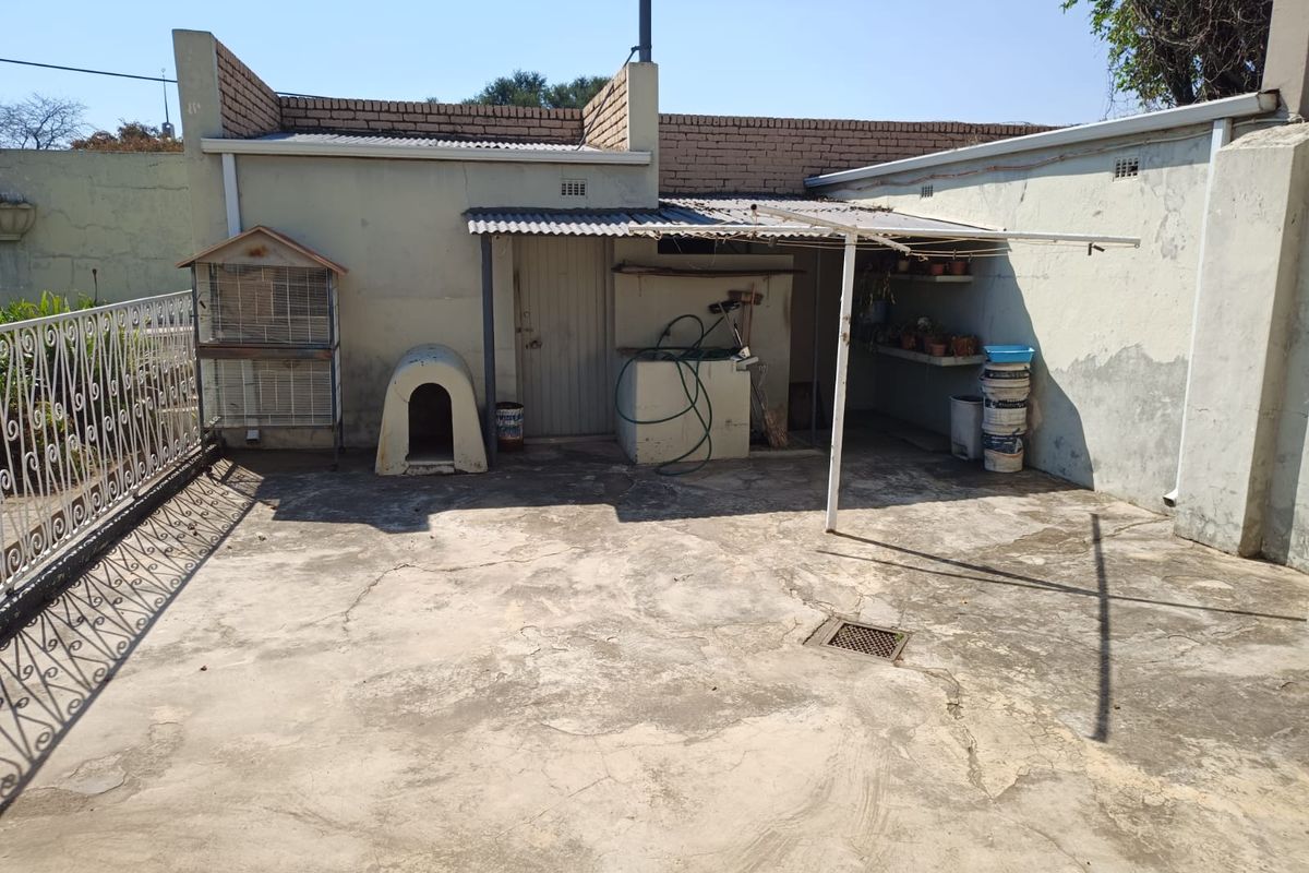 A rear exterior concrete patio area featuring a covered section with corrugated roofing, masonry walls, and a drainage system.