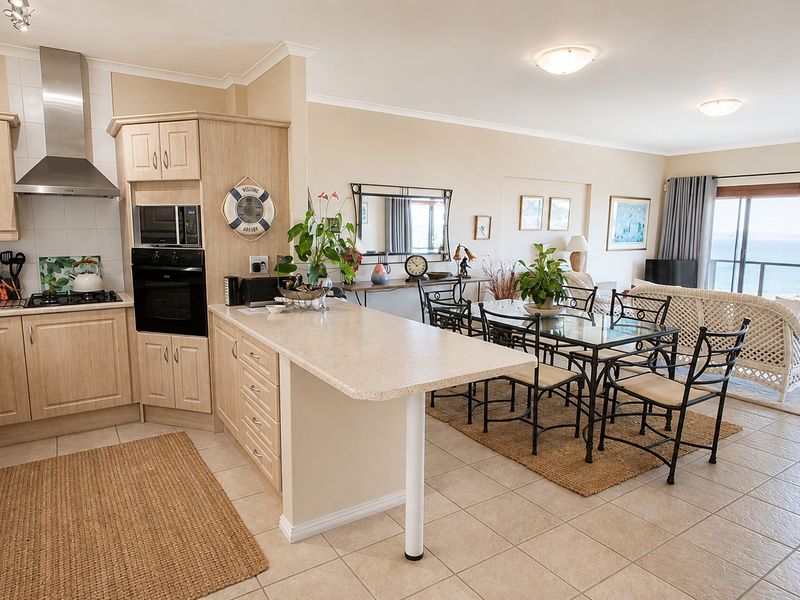 Open-plan kitchen and dining area featuring light wood cabinetry, integrated appliances, tiled flooring, and direct access to a balcony with ocean views.
