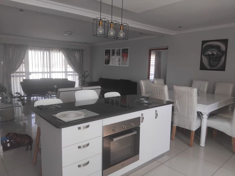 Modern open-plan living area featuring a central kitchen island with built-in oven and hob, flowing into a dining and lounge space with tiled floors.