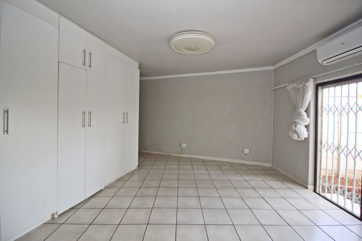 Spacious bedroom featuring neutral tiled flooring, extensive built-in storage cupboards, an air conditioning unit, and direct access to the exterior via a security gate.