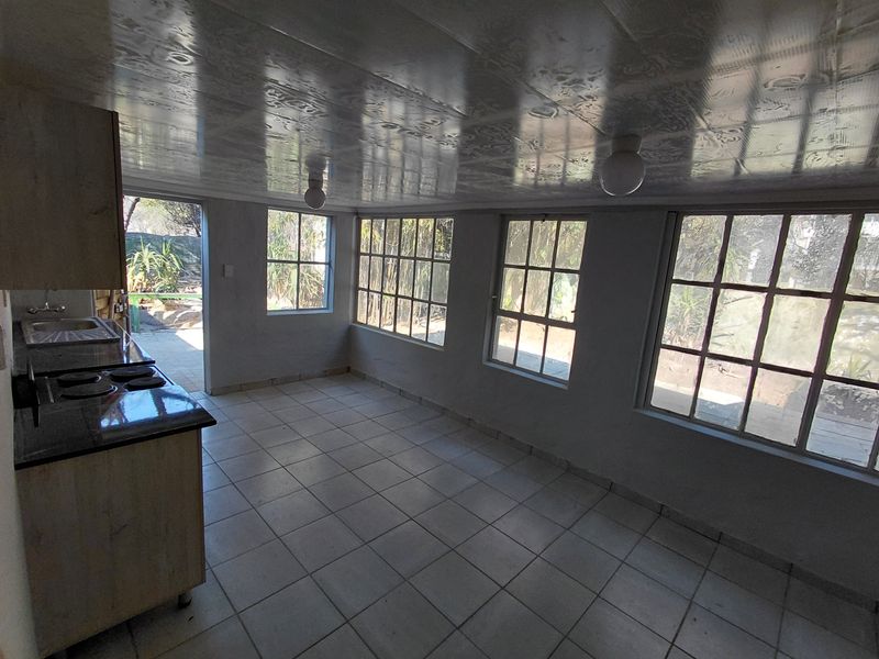 A bright, empty kitchen area featuring tiled floors, built-in cabinetry with a hob, and large grid-style windows overlooking the garden.