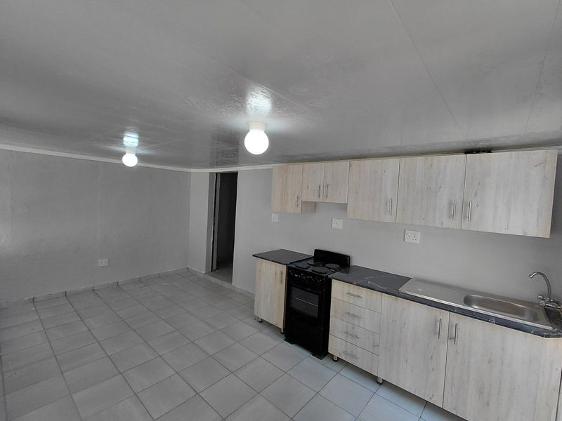 A functional, empty kitchen featuring light wood-look cabinetry, dark laminate countertops, a stainless steel sink, and a freestanding stove with tiled flooring.