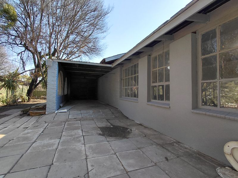 Rear exterior view of the cottage featuring a paved patio area, a covered carport, and large steel-framed windows along the side elevation.