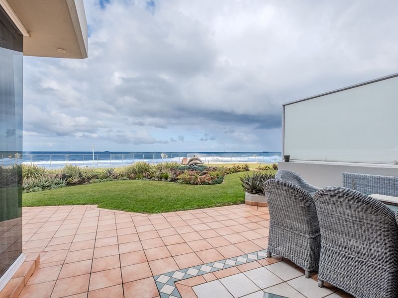 A spacious ground-floor patio featuring tiled flooring and a glass balustrade, overlooking a private garden with direct ocean views.