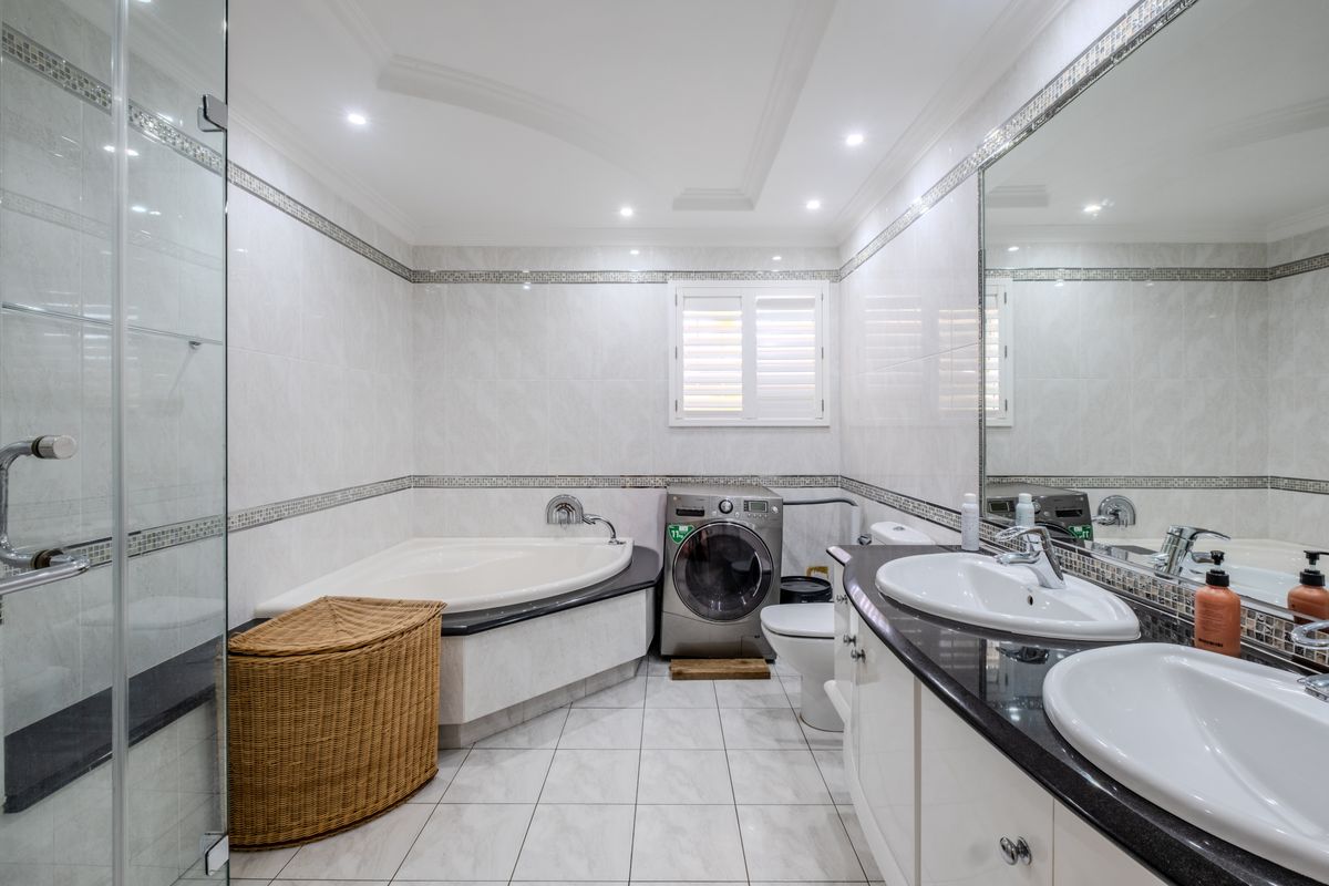 A well-appointed bathroom featuring a corner bathtub, double vanity with granite countertop, glass-enclosed shower, and integrated laundry facilities.