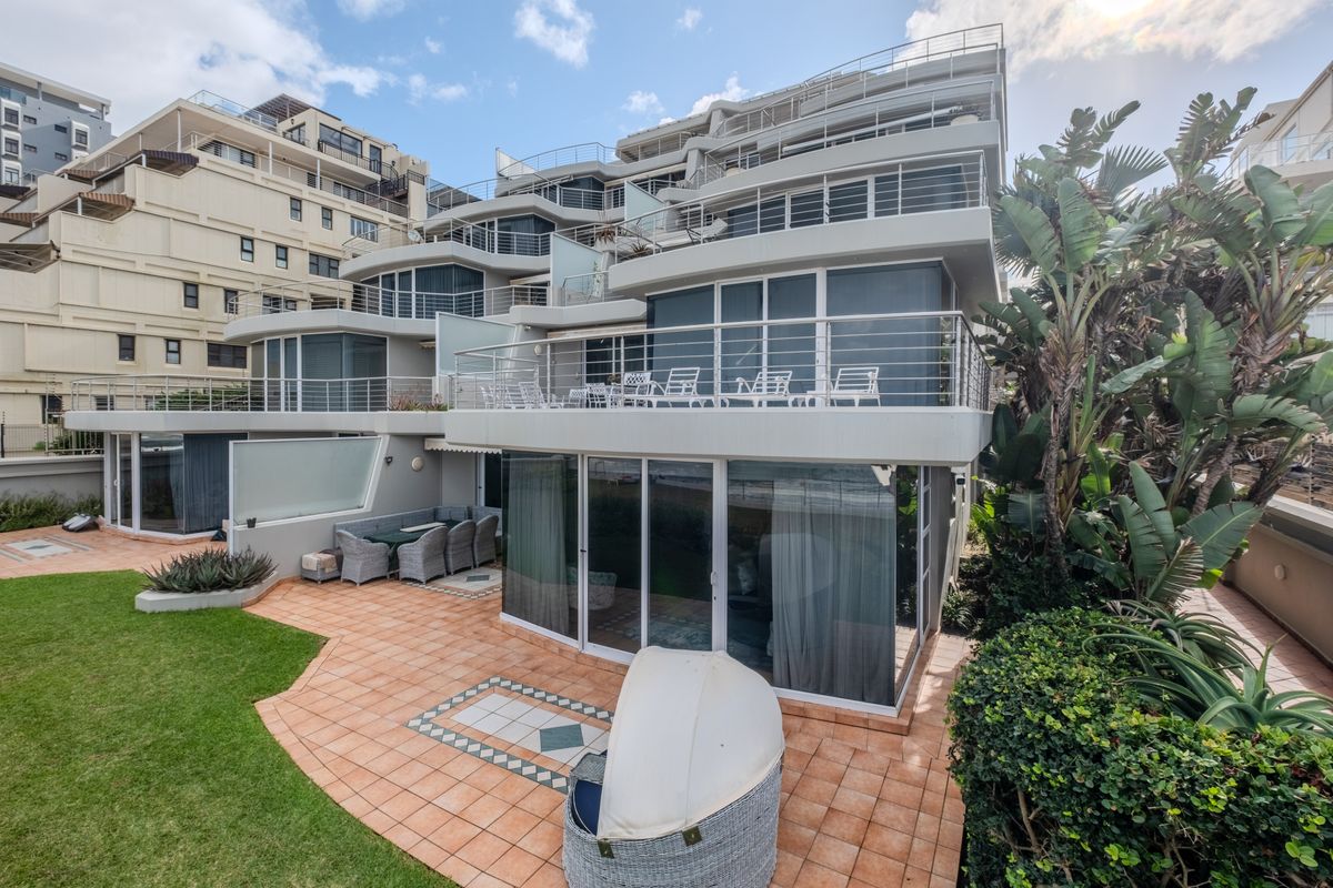 Rear view of a multi-story apartment complex featuring tiered balconies with stainless steel railings, sliding glass doors, and a paved patio area with garden.