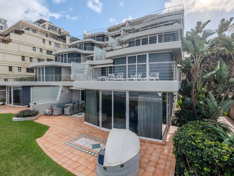Rear view of a multi-story apartment complex featuring tiered balconies with stainless steel railings, sliding glass doors, and a paved patio area with garden.