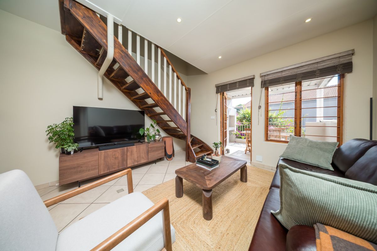 A bright living room featuring a prominent wooden staircase, tiled flooring, and direct access to an outdoor patio area through timber-framed doors.