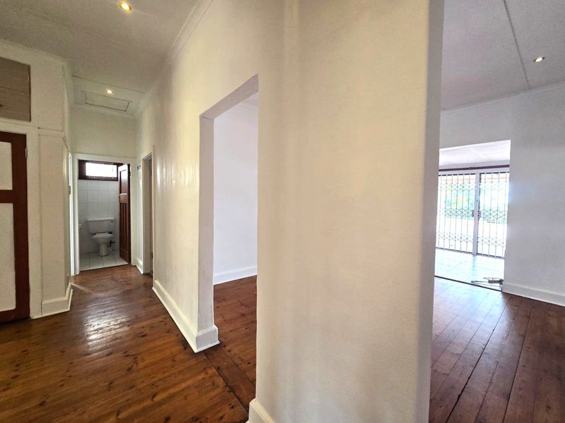 A bright, empty hallway featuring polished timber flooring, recessed downlighting, and access to adjacent rooms, including a bathroom and living area.