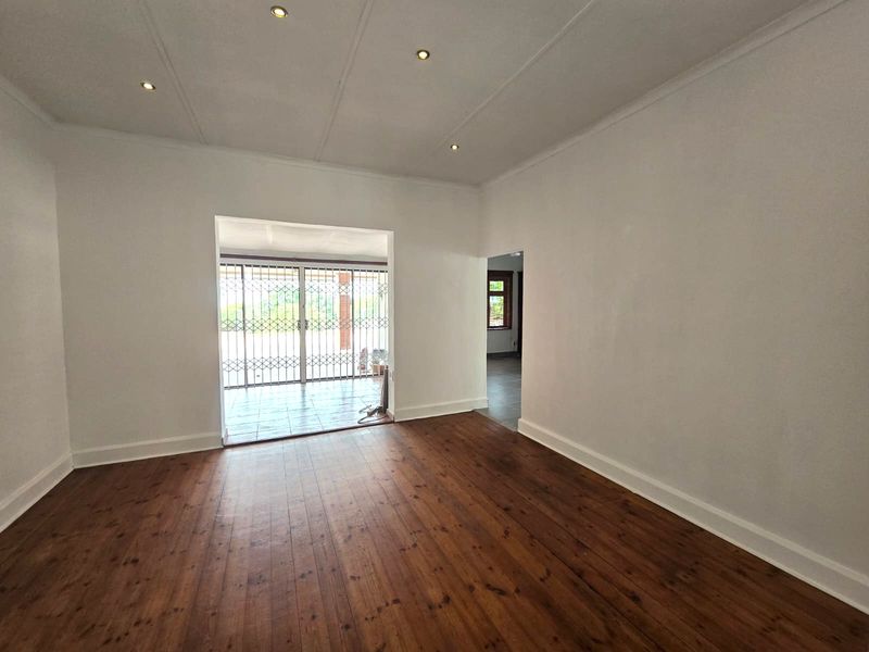 A bright, empty living room featuring polished timber flooring, recessed downlighting, and a security gate leading to an outdoor patio area.