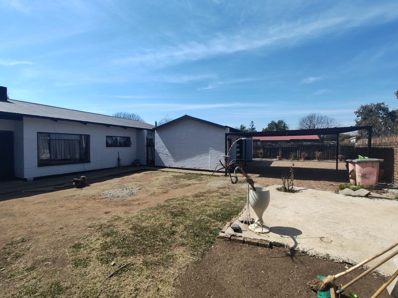 Rear exterior view of the property featuring a painted brick facade, a covered carport area, and a spacious backyard with potential for landscaping.