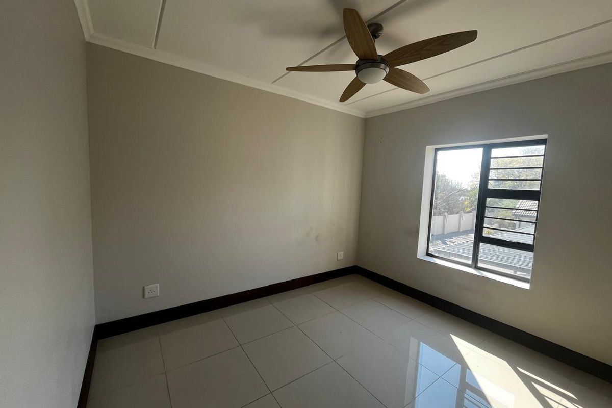 A bright, empty bedroom featuring neutral tiled flooring, a ceiling fan, and a window fitted with security bars.