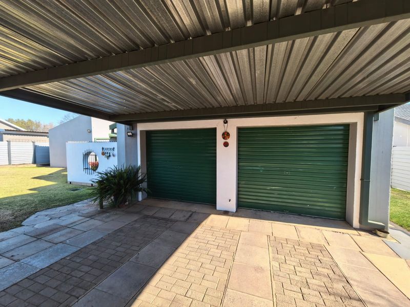 A double garage with green roller doors, covered by a corrugated metal carport, featuring a paved driveway and adjacent garden area.