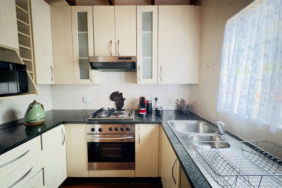 A functional kitchen featuring light-toned cabinetry, granite countertops, a gas hob with extractor, and a double basin sink under exposed timber ceiling beams.