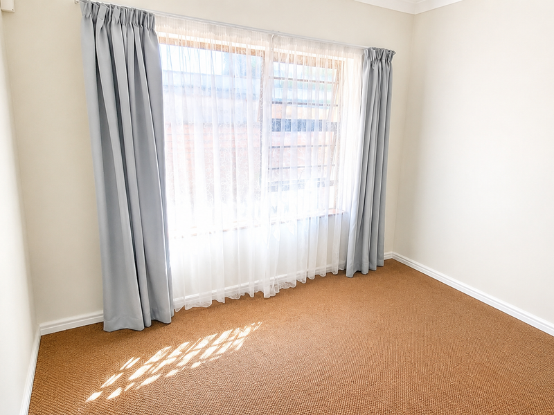 A bright, empty bedroom featuring neutral walls, carpeted flooring, and a large window fitted with security bars and curtains.