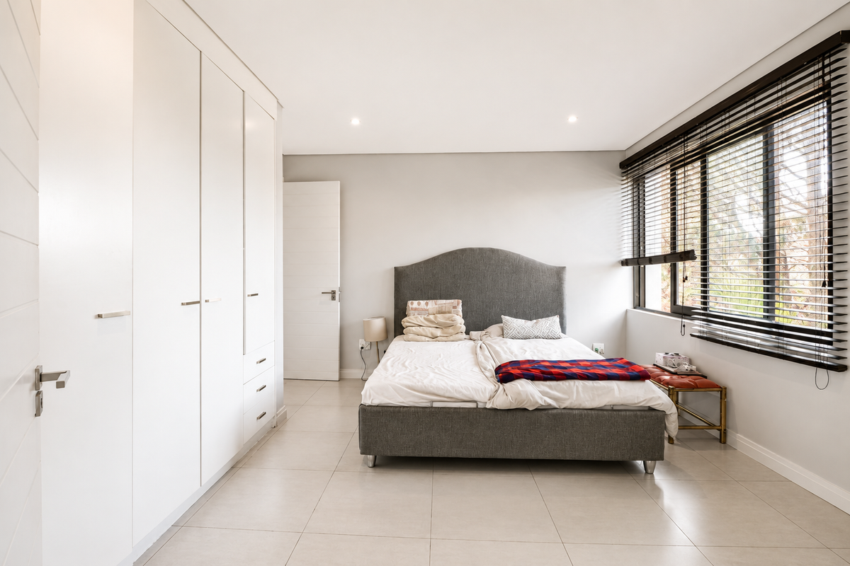 A bright and modern bedroom featuring neutral tiled flooring, extensive built-in cabinetry, and large windows fitted with venetian blinds.