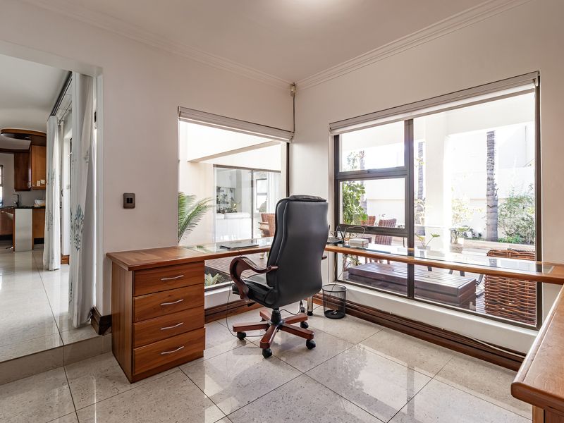 A bright home office featuring a custom built-in wooden desk, polished tile flooring, and large windows overlooking the outdoor patio area.
