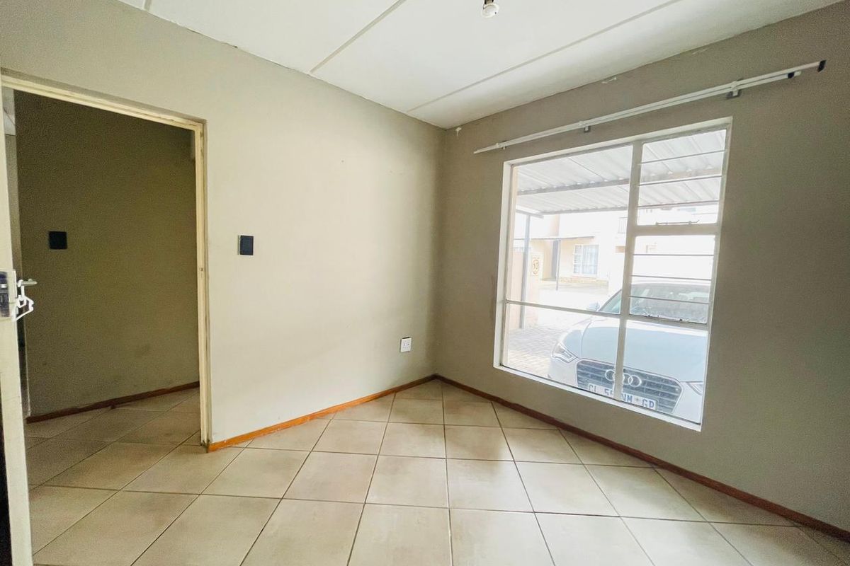 An empty living room featuring neutral tiled flooring, a large window with a curtain rail, and a simple ceiling light fixture.