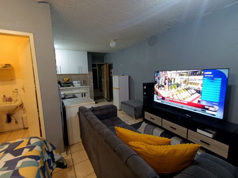 Open plan living area featuring tiled floors and an adjacent kitchen, with a view into a small bathroom with exposed plumbing.