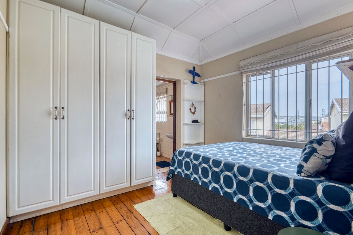 A bedroom featuring timber flooring, built-in cupboards, and a window with security bars, offering a view of the surrounding neighborhood.