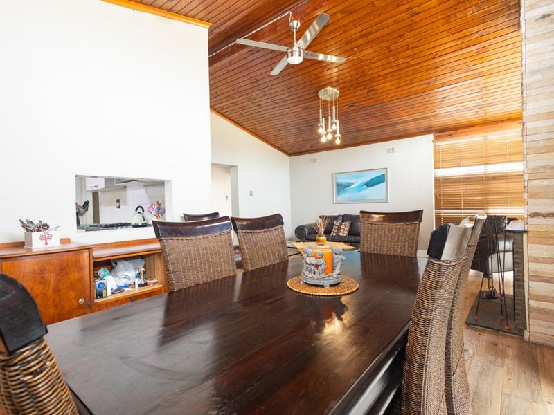 Open-plan dining and living area featuring a vaulted timber ceiling, ceiling fan, and a prominent stone-clad fireplace.