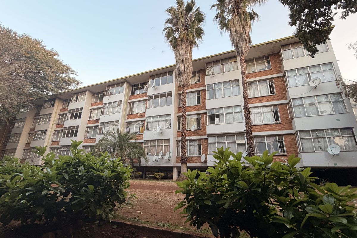 A multi-story residential apartment block featuring a brick facade and large windows, set within a landscaped garden area.