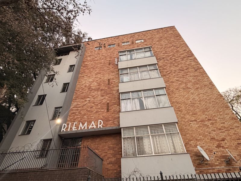 Low-angle view of the Riemar apartment building facade, featuring classic brick construction, multi-level window arrangements, and secure perimeter fencing.