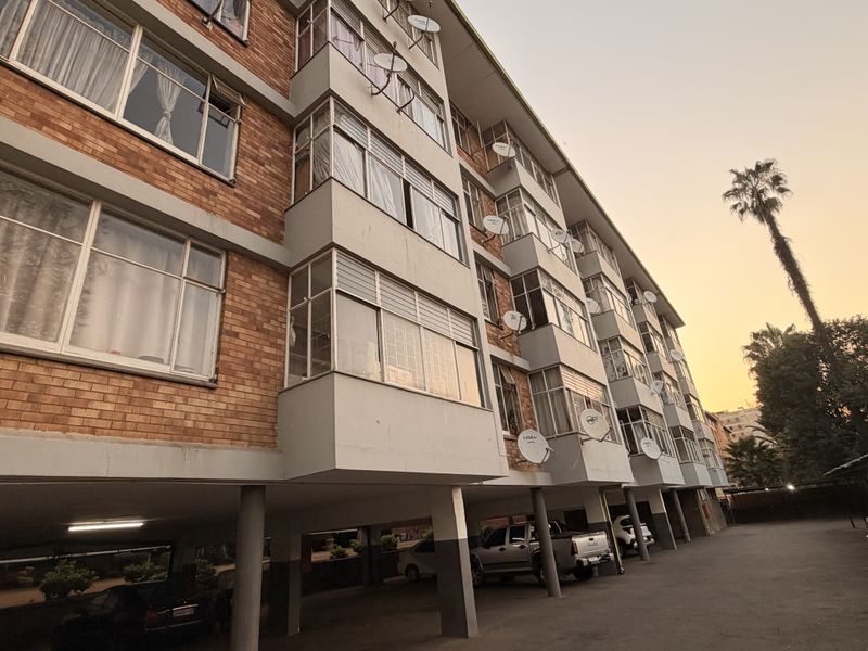 Exterior view of a multi-story apartment building featuring a brick facade, steel-framed windows, and a ground-level covered parking area.
