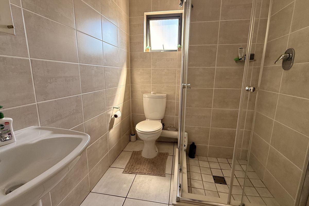 A functional bathroom featuring neutral ceramic wall and floor tiles, a pedestal washbasin, a toilet, and a glass-enclosed shower unit.