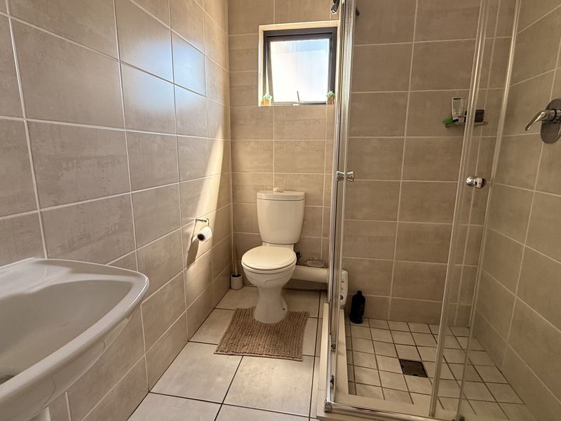 A functional bathroom featuring neutral ceramic wall and floor tiles, a pedestal washbasin, a toilet, and a glass-enclosed shower unit.