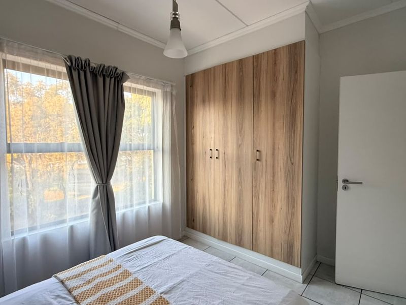 A well-lit bedroom featuring modern wood-finish built-in wardrobes, neutral tiled flooring, and a large window providing natural light.