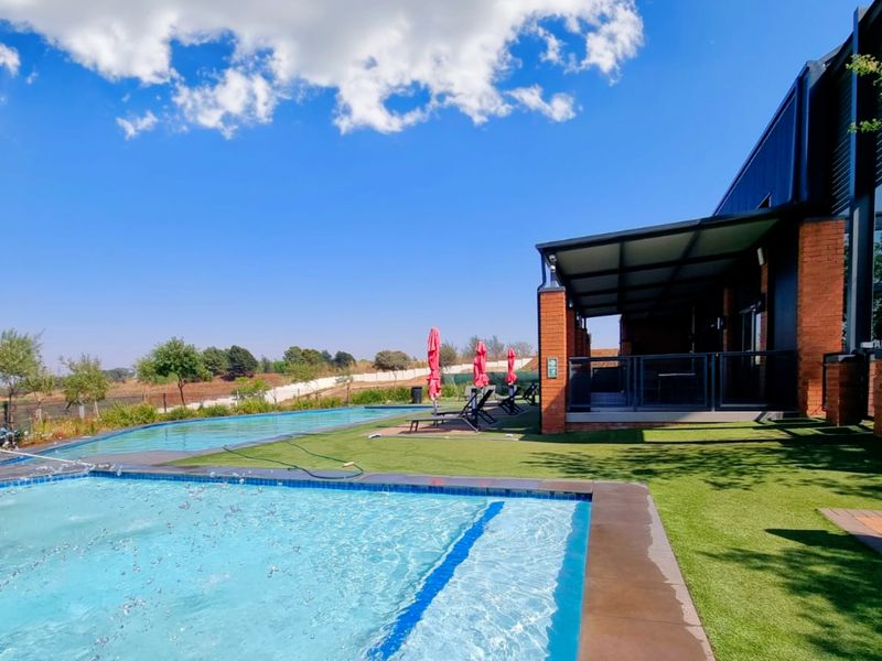 A well-maintained outdoor pool area featuring a swimming pool, artificial lawn, and a covered patio attached to a modern brick building.