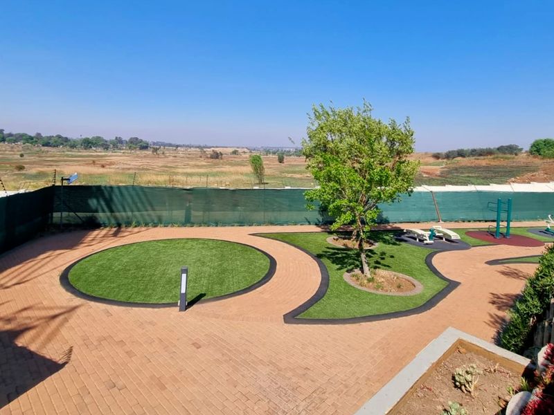 A well-maintained communal garden area featuring paved pathways, artificial lawn sections, outdoor gym equipment, and perimeter fencing with open landscape views.