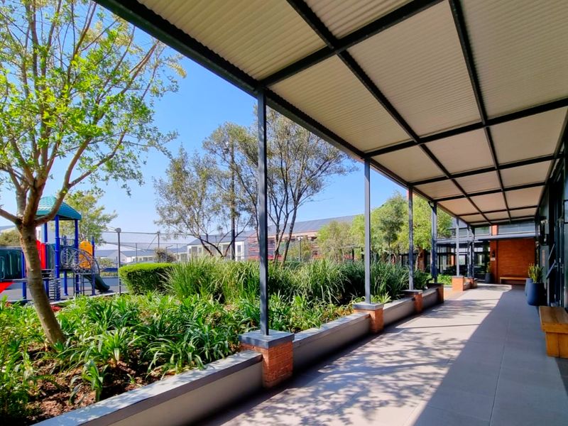 A covered outdoor patio area featuring tiled flooring, a corrugated metal roof, and a brick planter wall overlooking a landscaped garden and playground.