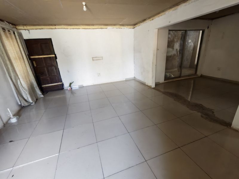 An empty open-plan living area featuring tiled flooring, a timber entrance door, and sliding glass doors, currently requiring significant cosmetic renovation and finishing.