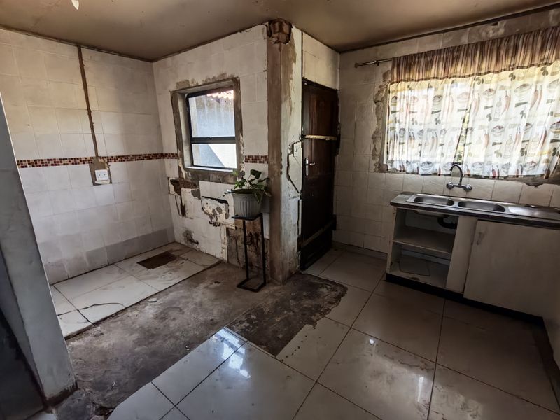 A kitchen in need of significant renovation, featuring tiled surfaces, a double sink, and visible damage to the walls and flooring.