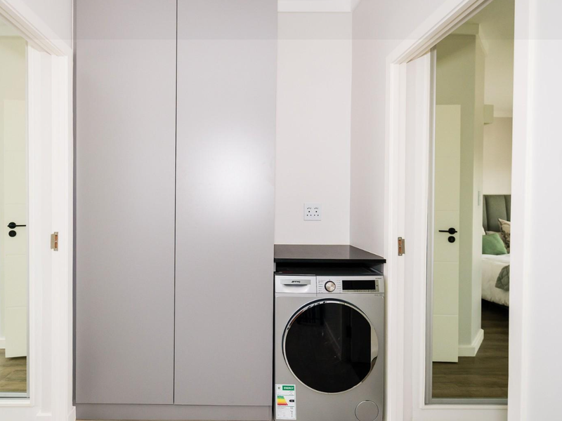 A compact, modern laundry nook featuring sleek built-in cabinetry and a fitted washing machine, conveniently located between two bedrooms.
