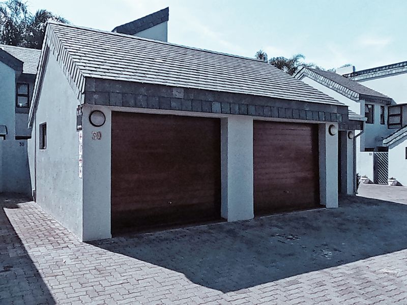 A double garage with wooden doors and a tiled roof, situated in a paved driveway area of the townhouse complex.