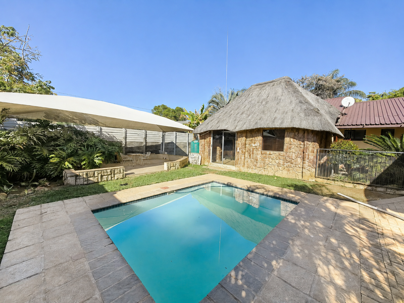 Outdoor pool area featuring a swimming pool, paved decking, a shade sail, and a unique thatched-roof structure with stone cladding.